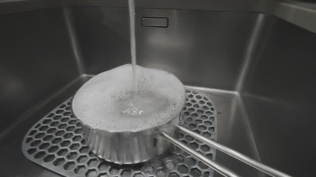 Running water from a faucet pouring into a metal pot with sudsy dish soap. Close up of cleaning cookware in a modern kitchen sink