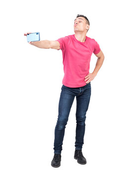 Full length of funny young guy in t shirt and jeans pouting lips and taking selfie on mobile phone with hand on waist