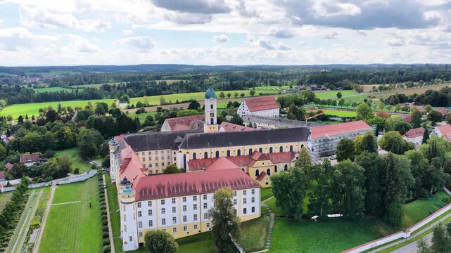 Malerische Luftaufnahme von Ochsenhausen mit der barocken Klosteranlage, eingebettet in die idyllische, gr&uuml;ne H&uuml;gellandschaft Oberschwabens unter blauem Sommerhimmel mit Wolken.