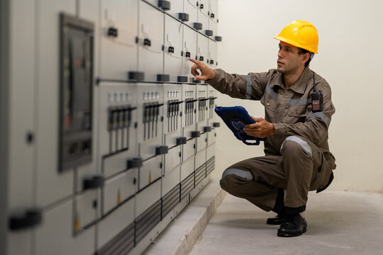 Industrial engineer inspecting electrical control panel in protective workwear, using tablet inside clean utility room with focused attention and caution