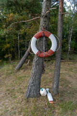 Red and white lifebuoy hangs from a pine tree with rope and marine buoys resting below. Outdoor lifesaving equipment represents water safety, rescue readiness, boating, emergency response and accident © Volha