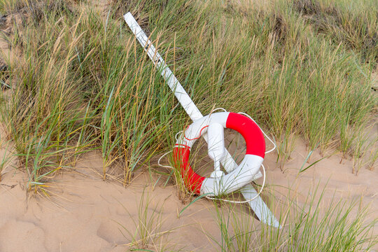 Red and white lifebuoy attached to rescue equipment lies among tall beach grass on a sandy coastal dune. Visible lifesaving gear represents water safety, emergency preparedness, rescue service and acc