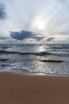 Rolling sea waves reach an empty sandy beach while filtered sunlight reflects across the water beneath dramatic clouds. Atmospheric coastal landscape for seaside travel, vacation, weather, nature and 