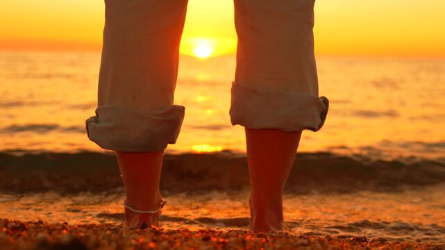 Summer vacation legs standing on golden coastline, soaking in vibrant sunset waves while relaxing outdoors. Fixed low angle long shot.