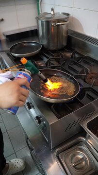 Chef using blowtorch to flambe and sear beef in frying pan on gas stove in commercial kitchen.