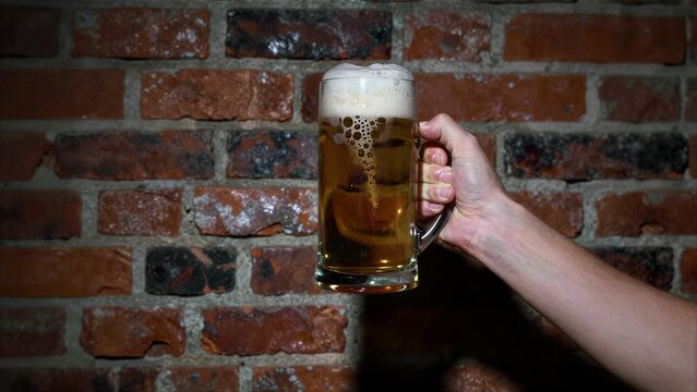 Hand holds beer mug against rustic brick wall, golden liquid with frothy head.