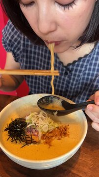 Happy food blogger young asian woman eating hot ramen soup noodle bowl inside vibrant traditional japanese restaurant enjoys authentic dish during lunch break for social content with calm joyful