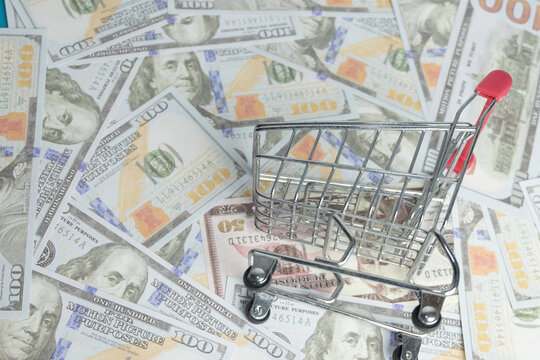 Miniature shopping cart on scattered US dollar bills. Concept of inflation, rising cost of living, expensive grocery shopping, purchasing power, e-commerce business and retail consumerism.