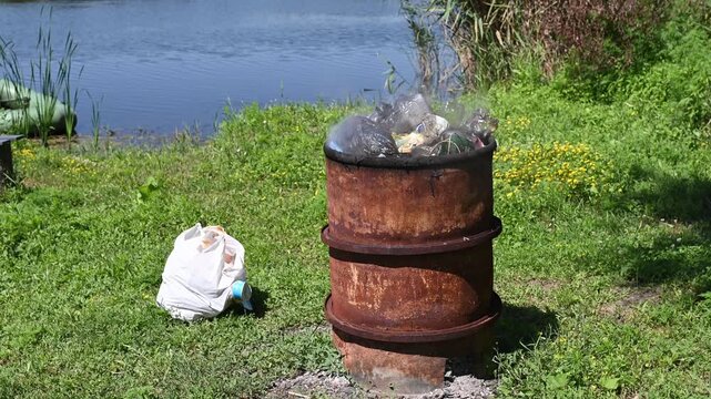Burning unsorted plastic waste in a metal barrel outdoors. Air and water pollution, environmental damage, toxicity. Environmental concept, illegal waste dumping, improper waste disposal.