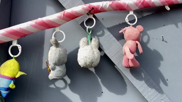 Small plush toys hanging from a candy cane pole against a gray wooden wall in bright sunlight. Playful background, cute stuffed animals, outdoor shadow, festive theme, and decorative toys.
