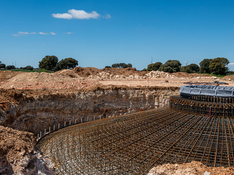 An expansive construction site photograph shows a deep, circular excavation pit for a wind turbine foundation, featuring an intricate, massive steel rebar reinforcement cage installed at the bottom, s