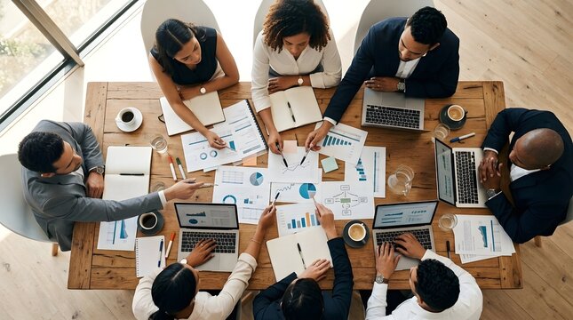 Multiple individuals in business attire are seated at a large wooden conference table  examining printed graphs  using laptops  and taking notes on paper pads