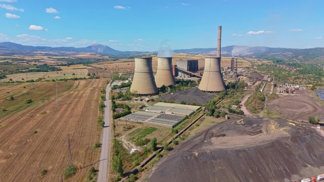 Thermal power plant dominates rural terrain, its cooling towers and chimney standing above fields. Steam drifts from plant as coal mounds and service roads mark ongoing energy production.