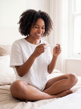 Pregnancy test positive black woman happy result motherhood fertility bedroom sits cross legged on a bright bed and smiles at her home test with relief for family planning and prenatal campaign use