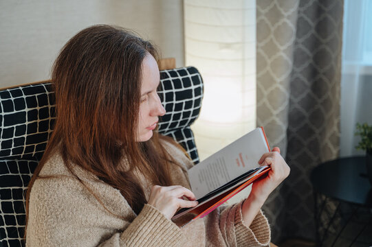 Woman wrapped in a soft warm throw blanket sitting in an armchair while reading an interesting book, cozy autumn lifestyle