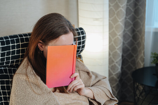 Young woman sitting in a cozy armchair covering her face with an open book, relaxed home reading and leisure concept