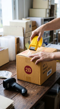 Hands sealing a cardboard box with tape dispenser on a wooden desk with shipping labels and scanner, symbolizing logistics and delivery process