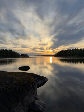 A serene sunset casts a striking golden glow through dramatic clouds, reflecting perfectly on a calm forest lake framed by dark, rocky shores.in Finland.