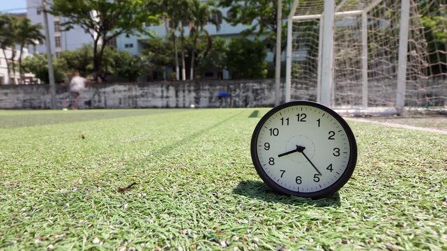 Abnormally fast moving clock hands spinning in selective focus on green artificial turf. A blurred ghosty teenager repeatedly kicks footballs into soccer goal net while trainer guides him