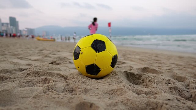 Orbiting shot of yellow soccer ball resting on sand as sea waves lap shore. Blurred background, incidental young girl in sprints toward water alongside few distant beachgoers under soft dusk sky.