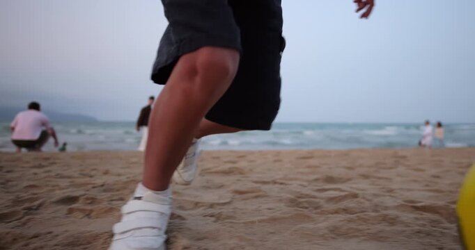 Youthful energy concept with close-up of boy's feet in white sneakers kicking bright yellow soccer ball on sandy beach during evening. In blurred background, incidental beachgoers stroll by
