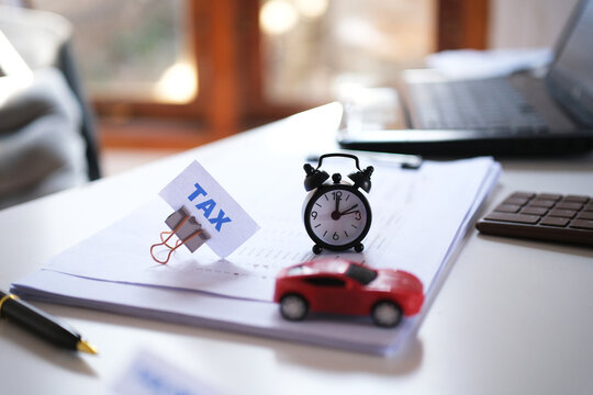 A sign reading "TAX" held by a clip, a toy car, and an alarm clock on a desk. Vehicle tax payment deadline. Financial planning, tax payment budget.