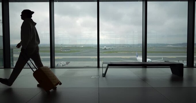 Silhouetted male traveler strolling at relaxed pace across spacious airport terminal hall while pulling trolley suitcase behind him. Stress-free travel lifestyle or successful flight connection