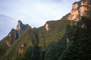 Rugged karst mountain summits blanketed in thick green foliage catch the golden rays of the sun. A solitary modern utility pylon stands on a steep ridge. © Sergej