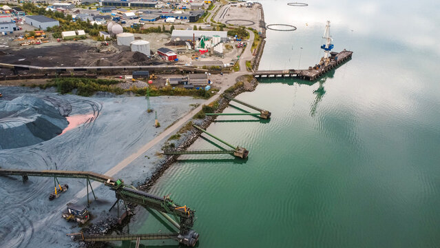 High-angle drone shot showing industrial loading conveyors, a dock harbor crane, and shipping facilities by turquoise water. Ideal asset for bulk freight, mining, and supply chain media.