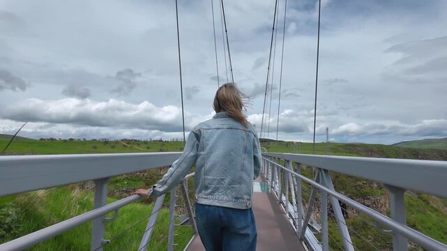 Young woman in denim jacket walking on modern suspension bridge with glass floor, enjoying the scenic view of the green Dashbash canyon on a cloudy day and feeling freedom