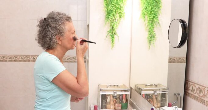 Mature woman applying blush to her cheeks with a makeup brush while looking into a bathroom mirror.