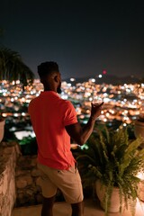 Obraz premium Man on terrace holding a wine glass overlooking city lights at night for lifestyle background and evening relaxation banner