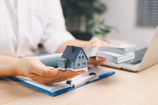 Person holding a miniature house and bundled banknotes beside loan documents and a laptop symbolizes property purchase, mortgage funding, investment, home financing, capital, and ownership.