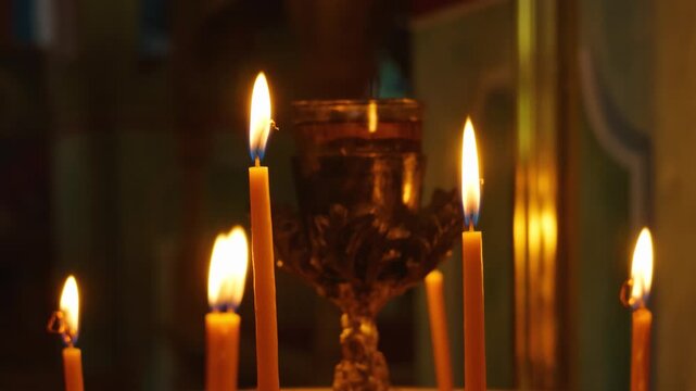 Close-up of several lit candles burning in a dimly lit church interior, with a metal chalice in the background, Tsagkarada, Pelion, Greece