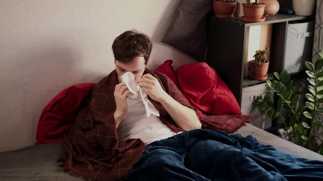 A sick man is sitting on a bed in a bedroom with a blanket over him. Young guy sneezing into a tissue. Diseases, viruses, and disorders
