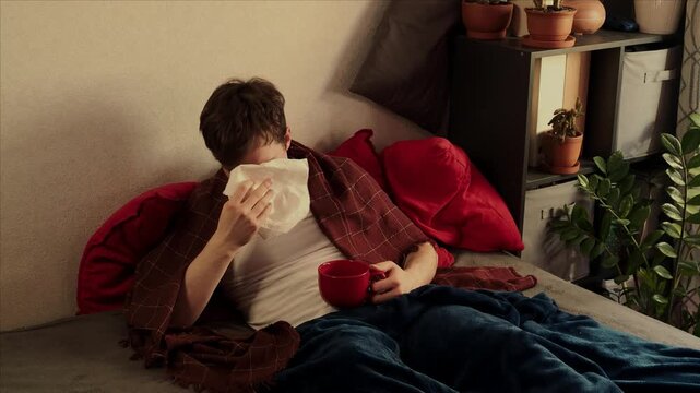 A sick man is sitting on a bed in a bedroom with a blanket over him. Young guy sneezing into a tissue. Diseases, viruses, and disorders
