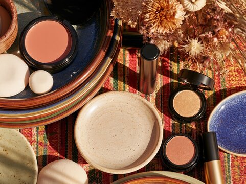 Cosmetics and ceramic dishes arranged on colorful woven textile with dried flowers, makeup compacts, brushes, and beauty sponges in an overhead flat lay composition