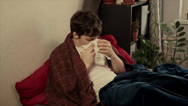 Sick guy with cold, flu or virus illness with cough and running nose lying in bed and using tissues. Sneezing ill guy at home. Man and health