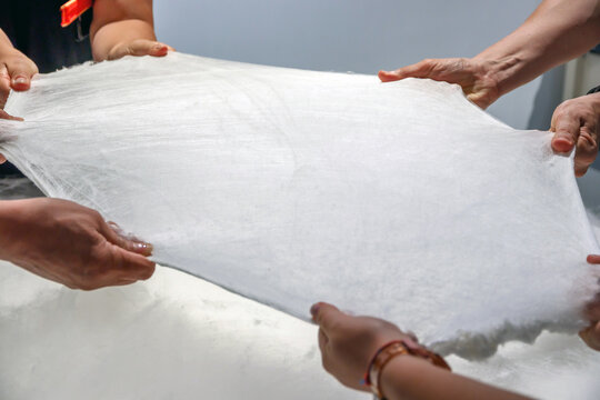 Multiple hands pull and stretch a fragile white sheet of raw silk in a hands-on chinese luxury workshop, showcasing traditional artisan technique