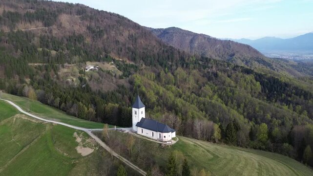 Aerial view of Sts. Primus and Felician Church in Jamnik, Slovenia, standing on a green hill surrounded by spring forest, with a scenic valley and snow-capped Julian Alps in the background.
