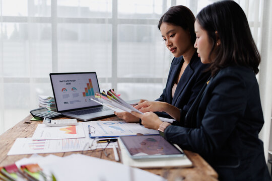 Two businesswomen collaborating on financial data analysis, reviewing charts and reports with a laptop and digital tablet, discussing business strategy and market growth