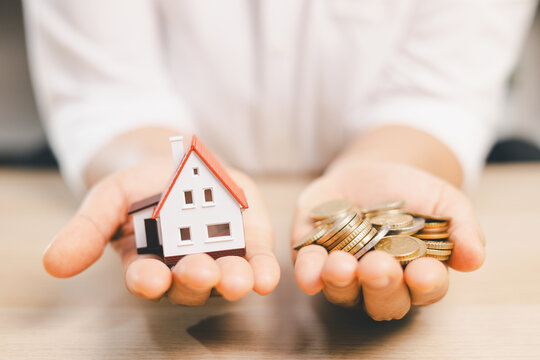 Hands holding a miniature house and coins symbolize home investment, mortgage affordability, property finance, savings, ownership, financial planning, and balancing housing costs with available money.