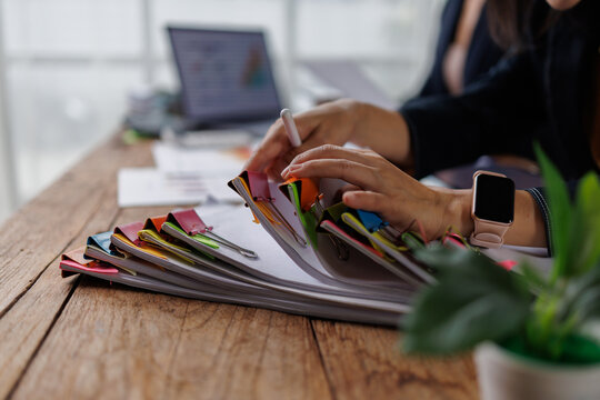 Businesswoman hands reviewing corporate financial documents and reports on rustic wooden desk, managing important office paperwork with binder clips and smart device