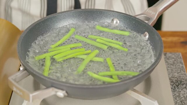 Crisp fresh green beans being blanched in boiling water in a shallow frying pan. Real-time footage showing vibrant green colors, gentle bubbling water and rising steam.