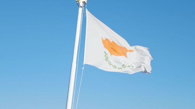 The national flag of Cyprus fluttering on a flagpole against a clear blue sky. The white flag features an orange map of the island above two olive branches