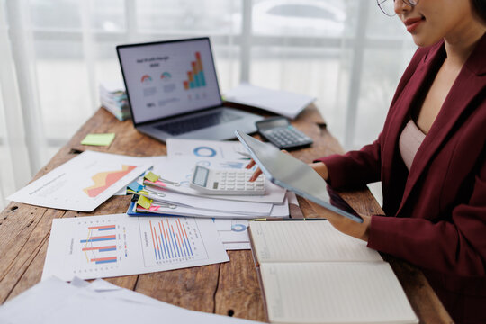Businesswoman at a cluttered desk analyzing financial data on tablet and calculator, reviewing charts and reports while multitasking through budgeting, planning and admin tasks