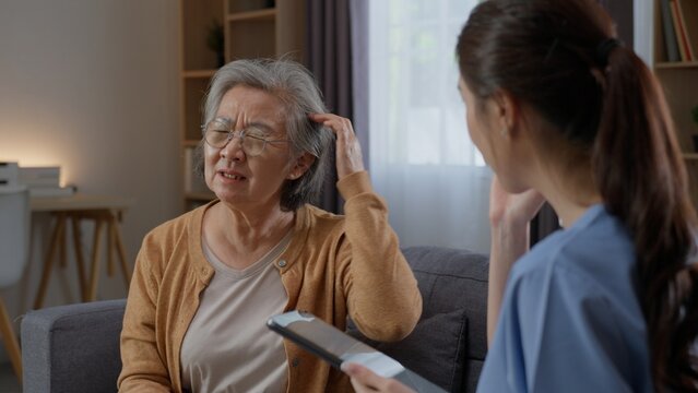 Elderly Asian woman touches her head while describing discomfort to a home care nurse, communicating a possible headache or pain symptom during a senior health assessment and consultation at home.