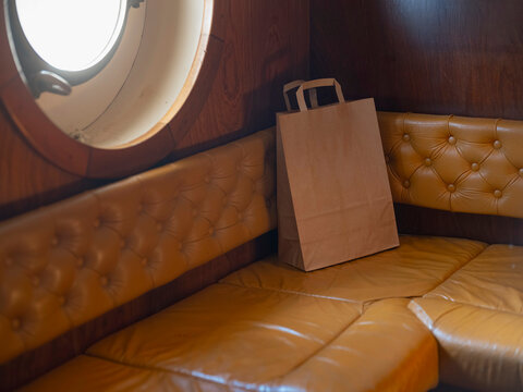 A blank kraft paper bag rests on a buttoned leather bench in a wood panelled ship's saloon beside a bright porthole, ready for branding.