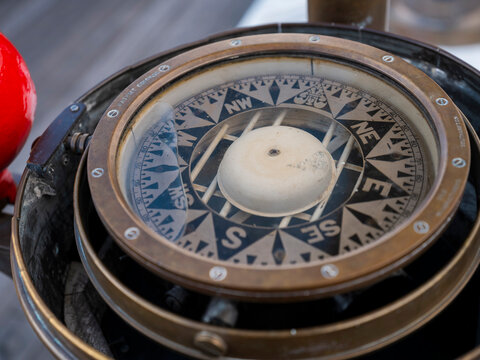 An antique brass ship's compass with its worn compass rose and red correcting sphere stands on the grey deck of a sailing ship.