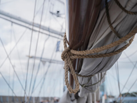 A rope loop and knot hold a furled sail against a varnished spar on a sailing ship under a soft grey overcast sky.